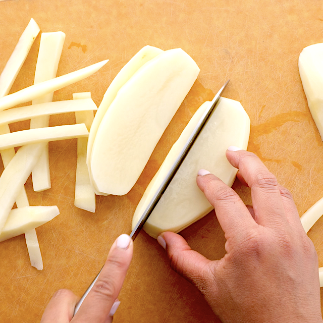 Close-up of hands slicing peeled potatoes into thin strips on a wooden cutting board.