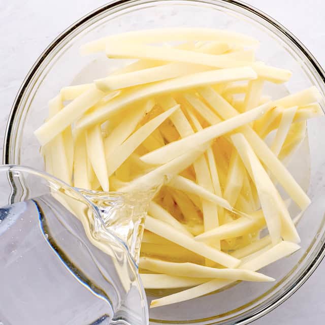 Water being poured over raw, cut potato sticks in a glass bowl.