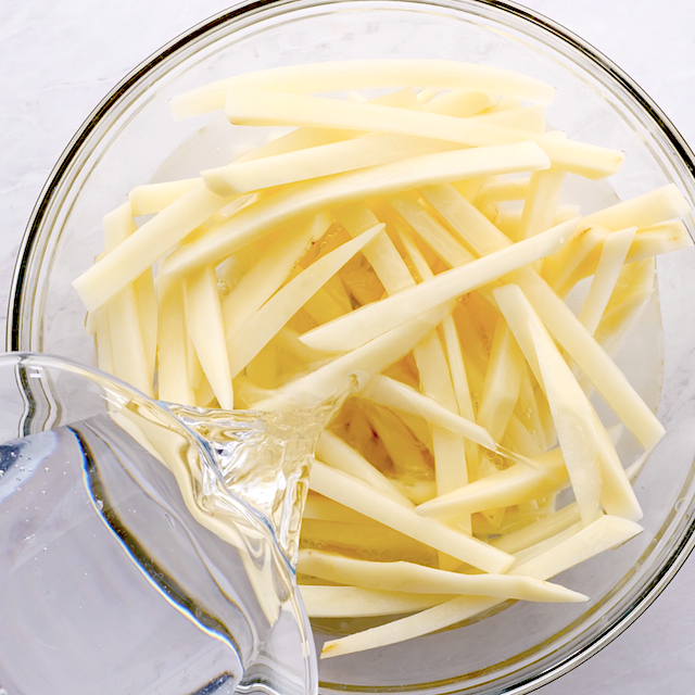 Water being poured over raw, cut potato sticks in a glass bowl.