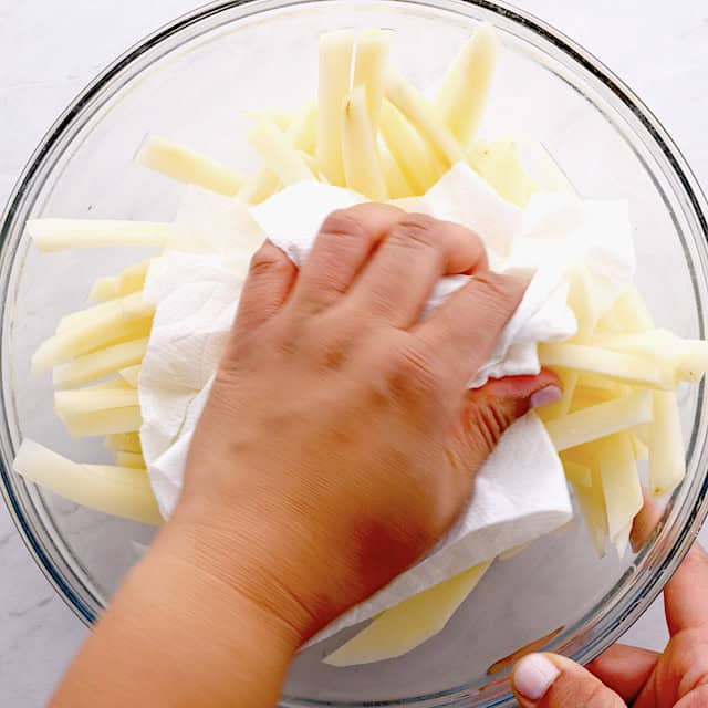 A person drying sliced potatoes with a paper towel in a glass bowl.