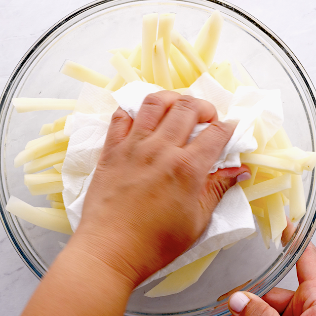 A person drying sliced potatoes with a paper towel in a glass bowl.