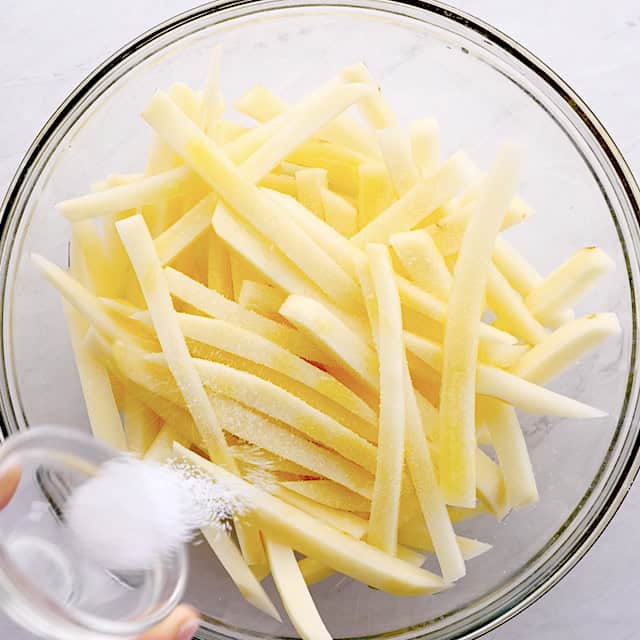 A person sprinkling salt over sliced potato strips in a glass bowl.