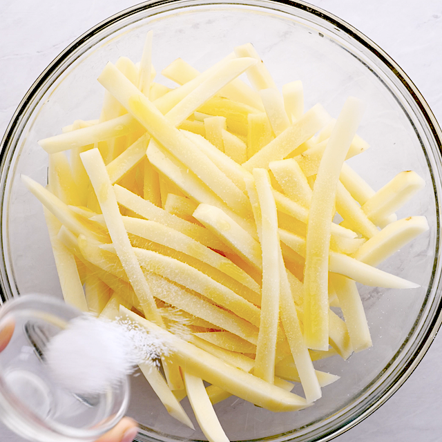 A person sprinkling salt over sliced potato strips in a glass bowl.