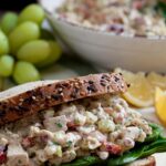 a healthy chicken salad sandwich with green grapes and a bowl of chicken salad in the background