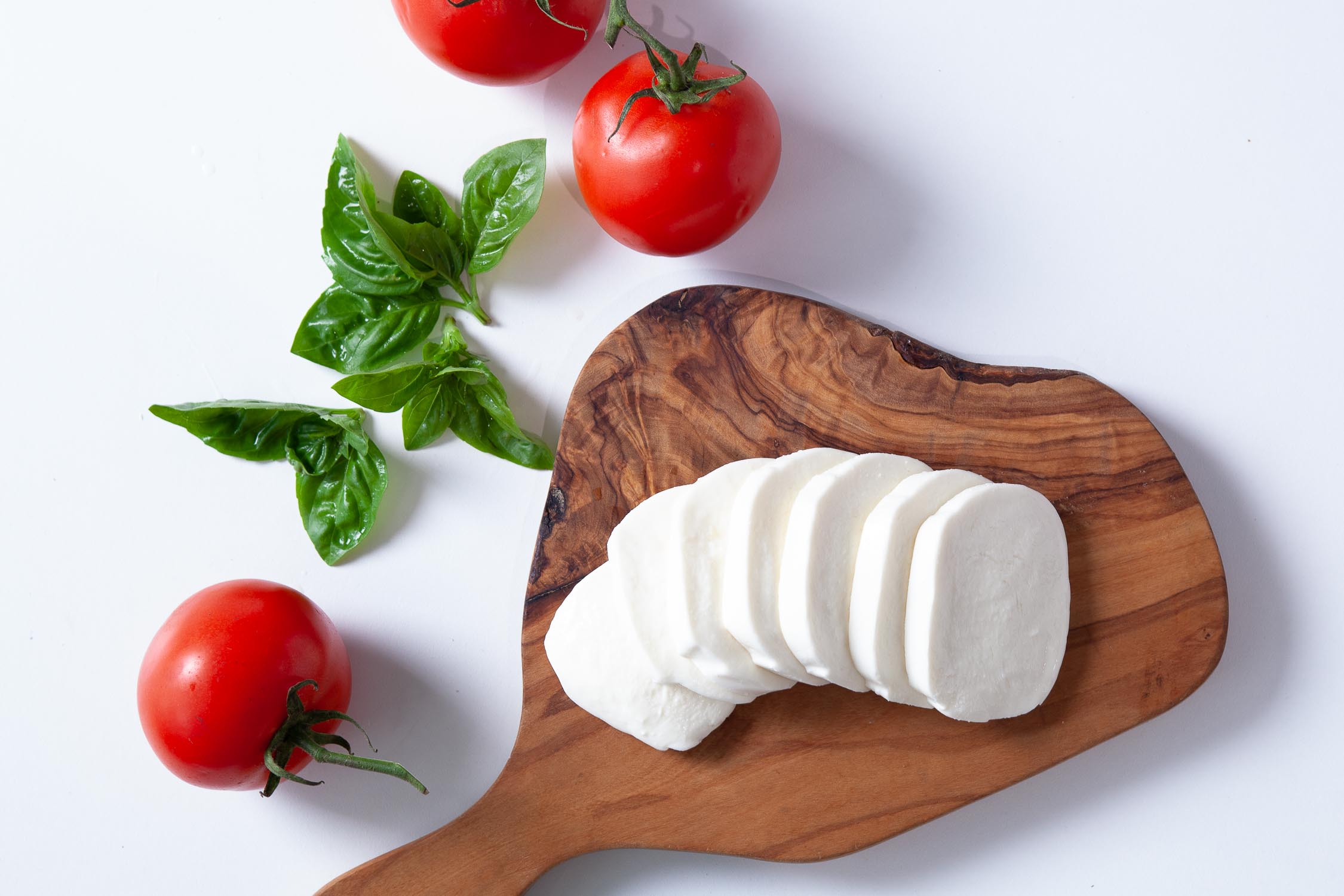 sliced fresh mozzarella on a wooden cutting board with tomatoes and basil on the side