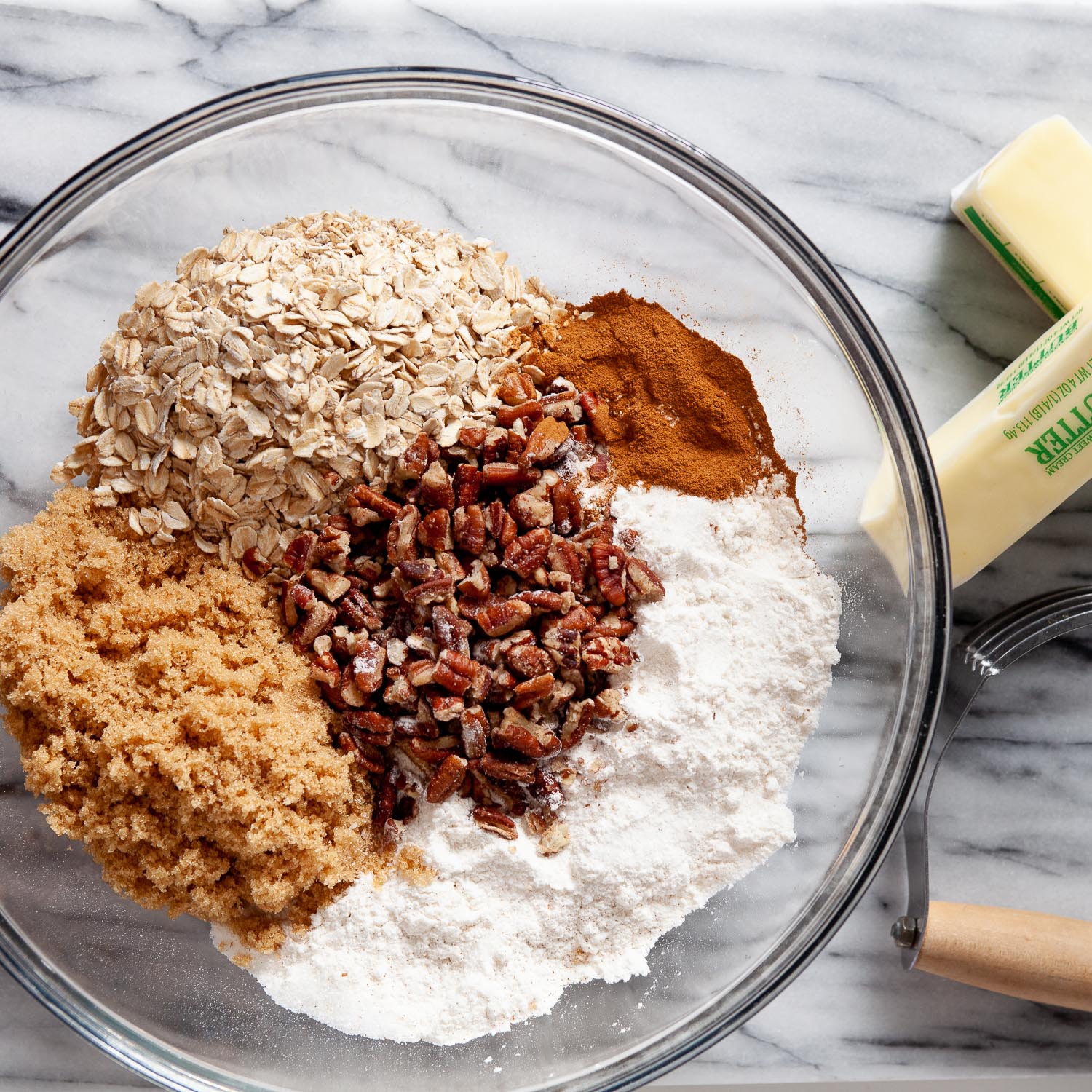 overhead image of a glass bowl full of oats, brown sugar, gluten free flour, chopped pecans, and cinnamon with two sticks of butter and a pastry cutter on the side