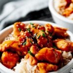 close up image of a white bowl full of teriyaki chicken on top of white rice topped with scallions and sesame seeds