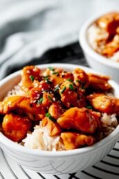 close up image of a white bowl full of teriyaki chicken on top of white rice topped with scallions and sesame seeds