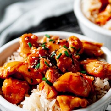 close up image of a white bowl full of teriyaki chicken on top of white rice topped with scallions and sesame seeds