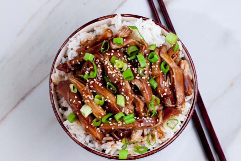 A bowl of rice topped with teriyaki chicken garnished with sesame seeds and chopped green onions. Chopsticks are placed beside the bowl on a marble surface.