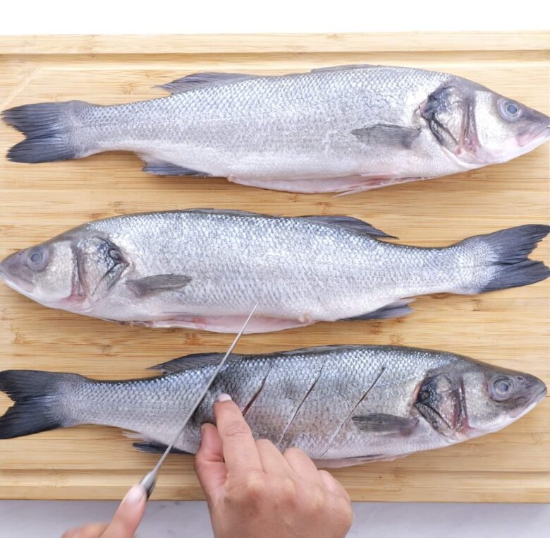 Branzino fish being scored with sharp knife on a cutting board.