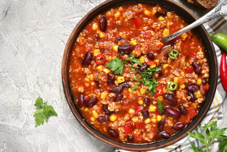 A bowl of hearty chili with ground meat, red kidney beans, corn, and diced tomatoes, garnished with fresh parsley and sliced chili peppers. A silver spoon rests in the bowl, set on a textured gray surface with a striped cloth underneath.
