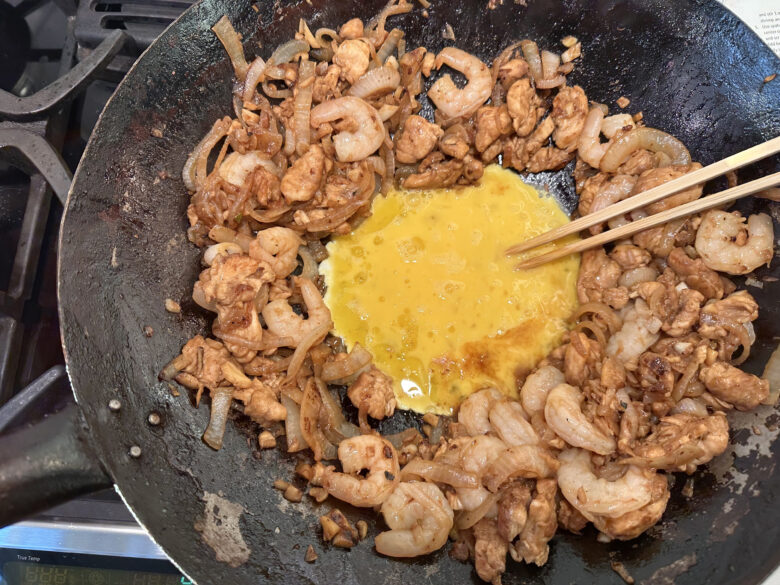 eggs being cracked in the well of a pan surrounded by pad thai ingredients