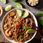 pad thai in a bowl topped with limes surrounded by bean sprouts, chili paste, lime wedges, and peanuts