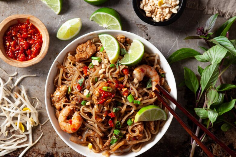 pad thai in a bowl topped with limes surrounded by bean sprouts, chili paste, lime wedges, and peanuts