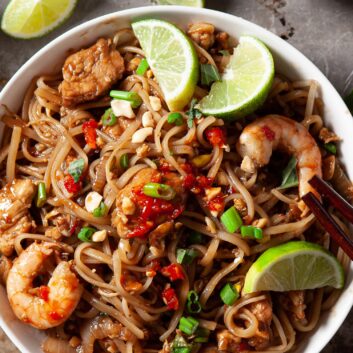 a bowl of pad thai with shrimp, chicken, and eggs topped with lime wedges and green onions