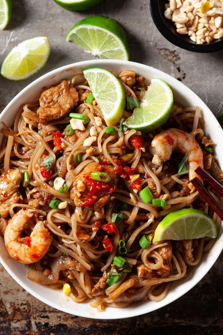 a bowl of pad thai with shrimp, chicken, and eggs topped with lime wedges and green onions