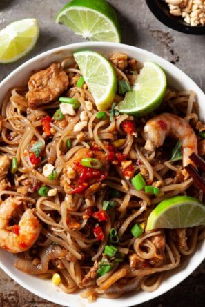 a bowl of pad thai with shrimp, chicken, and eggs topped with lime wedges and green onions