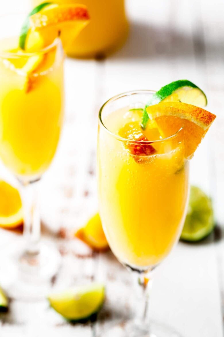 Two flutes of mimosas with slices of limes and oranges for garnish. 