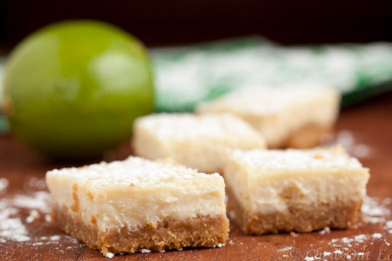 Four small, square lime bars with a crumbly base and creamy top layer are arranged on a wooden surface sprinkled with powdered sugar. A whole lime is partially visible in the background, adding a hint of green to the scene.