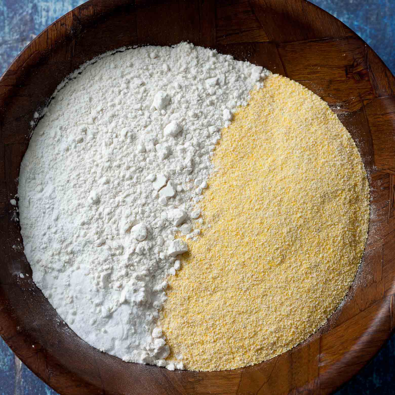 Flour and yellow cornmeal side by side in a wooden bowl, ready to be whisked together.