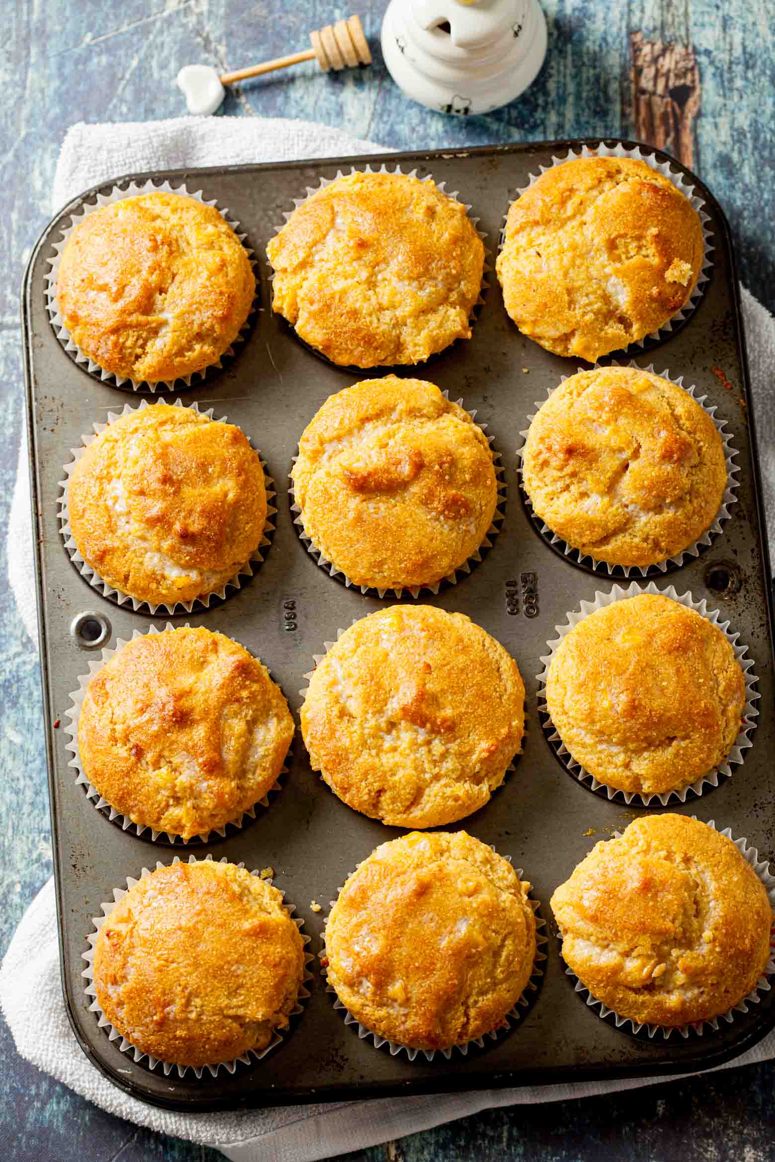 Twelve golden honey cornbread muffins in a muffin tin, with a honey dipper and a pot of honey in the background.