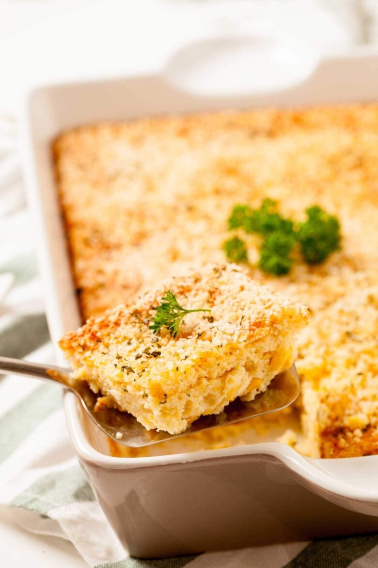 A piece of corn casserole on a serving spoon with the rest of the corn casserole in a baking dish in the background.