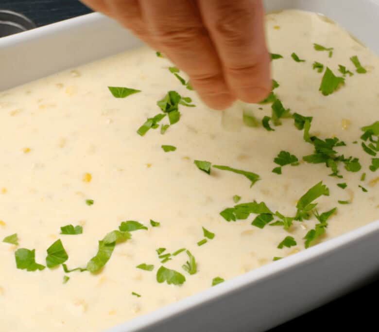 Baking dish with easy corn casserole batter with a person sprinkling fresh chopped parsley on top.