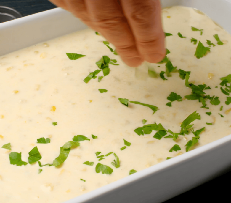 Baking dish with easy corn casserole batter with a person sprinkling fresh chopped parsley on top.