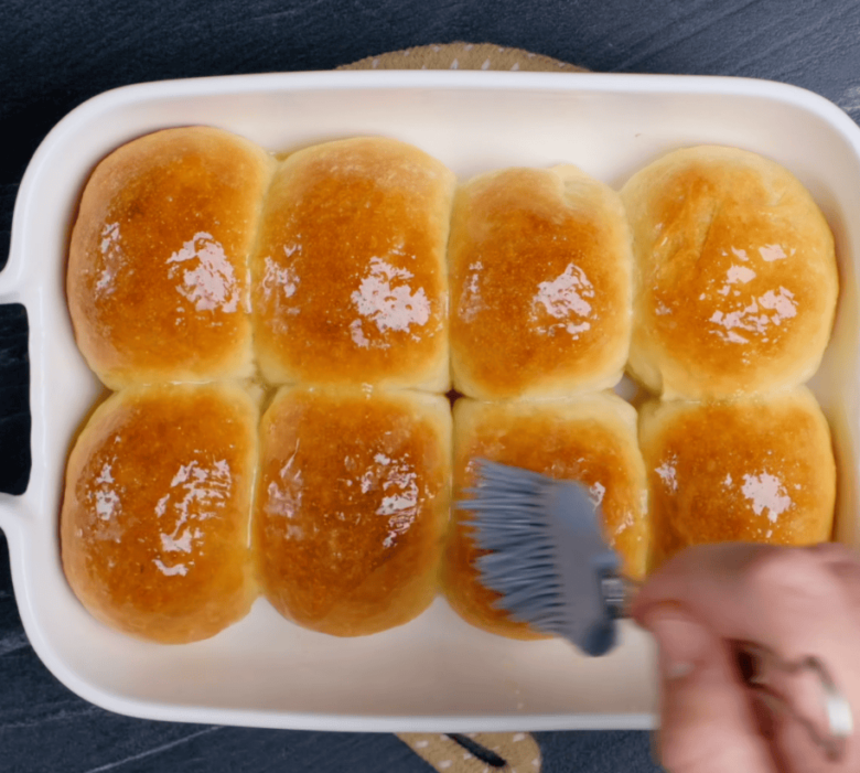Freshly baked easy dinner rolls being brushed with melted butter using a silicone pastry brush.