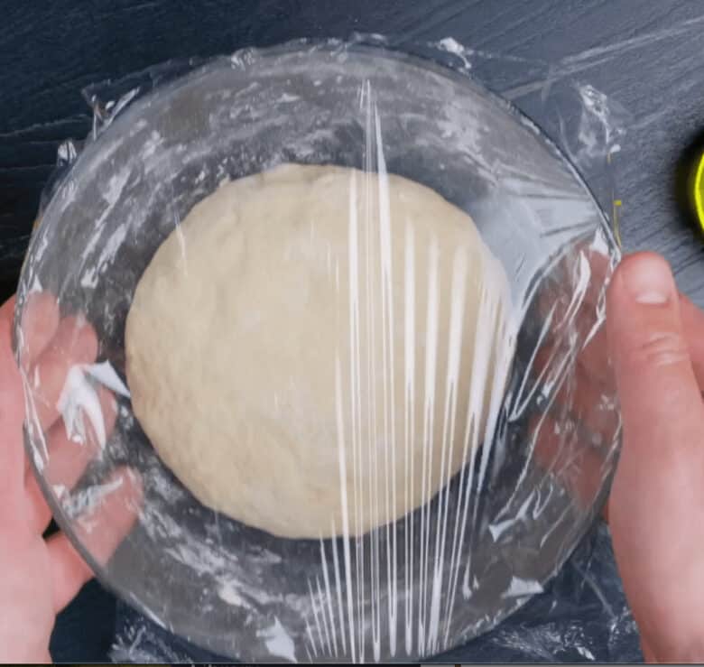 A bowl with kneaded dough covered in plastic wrap and being held by two hands.