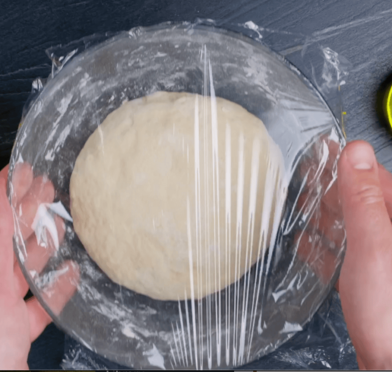 A bowl with kneaded dough covered in plastic wrap and being held by two hands. 