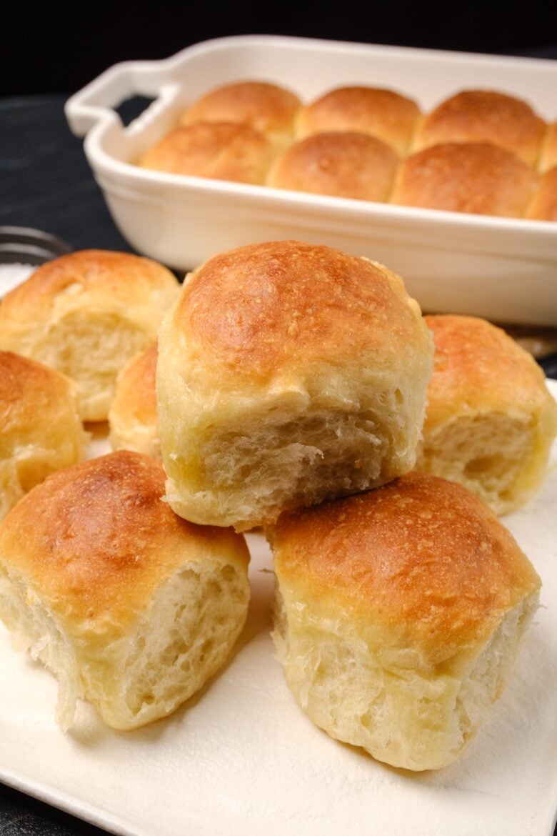 Soft and fluffy dinner rolls resting on a white plate with a baking dish with more dinner rolls in the background.