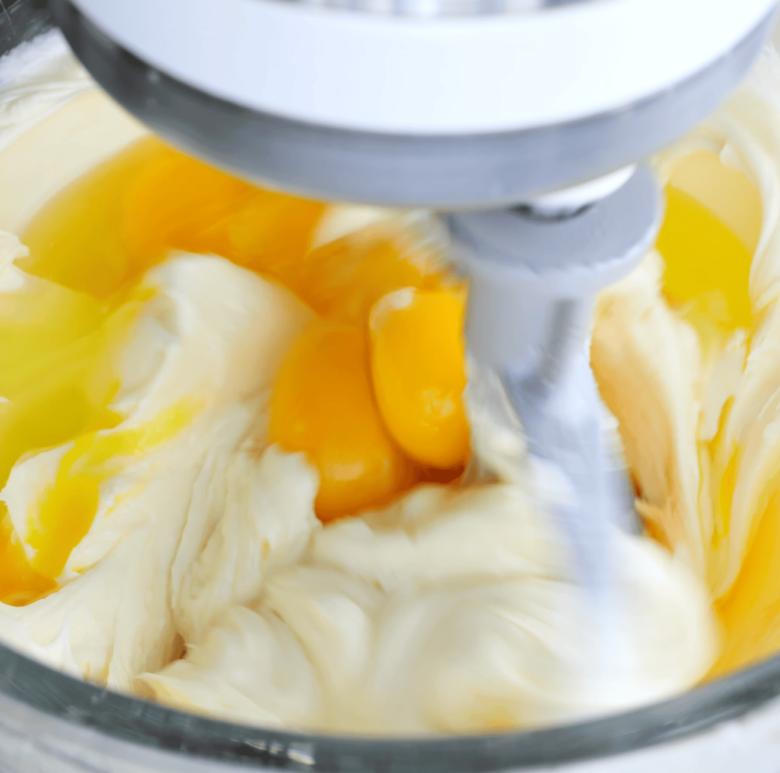Eggs and egg yolk being blended along with cream cheese in the bowl of a stand mixer.