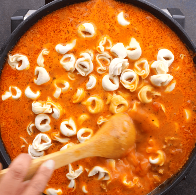 Frozen tortellini being stirred in a pan with tomato-based pasta sauce and other ingredients for Tuscan tortellini recipe.