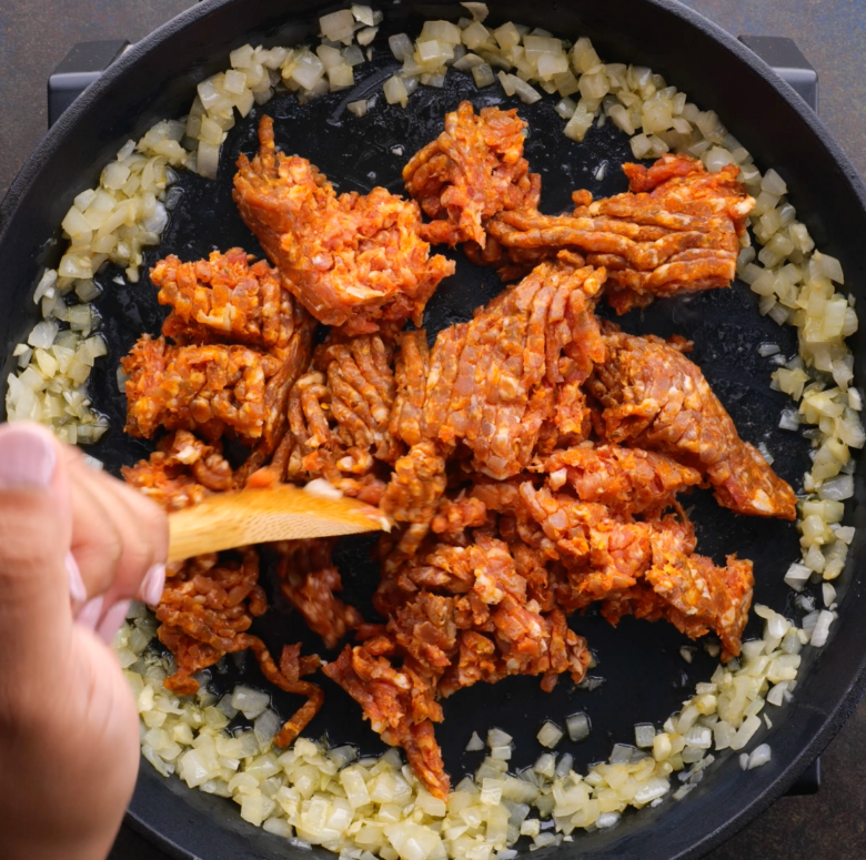 Bulk Italian sausage without casing in the center of a pan with sauteed onions and garlic towards the edges.