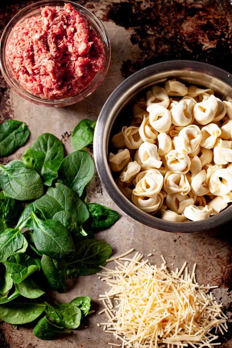 Overhead shot of ingredients for Tuscan tortellini recipe, including tortellini, baby spinach, and bulk Italian sauce without casings.
