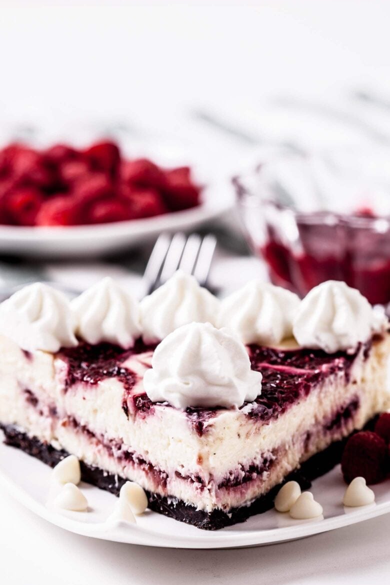 A closeup shot of a slice of raspberry cheesecake on a white plate with white chocolate chips around it.