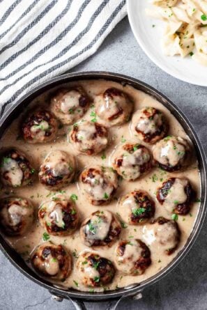 Overhead shot of easy Swedish meatballs recipe in a skillet with buttered noodles on a white plate in the corner.