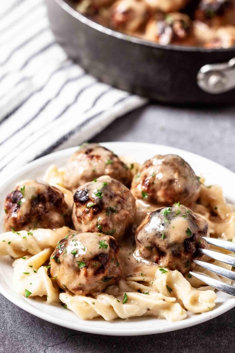 Closeup shot of Swedish meatballs in gravy over buttered noodles on a white plate with a fork.