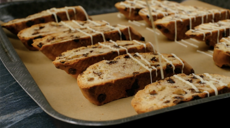 Baked almond biscotti being drizzled with melted white chocolate. 