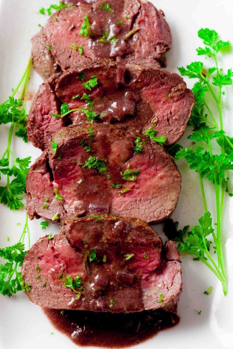 Sliced beef tenderloin roast on a serving plate with red wine saucce on top.