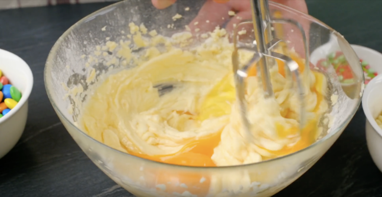 Eggs being beaten in a butter and sugar mixture with a hand whisk in a glass bowl.