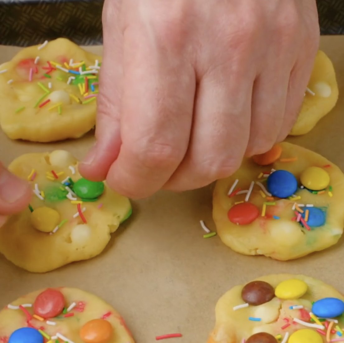 Extra M&Ms and sprinkles being added by hand on flattened M&M cookie dough.