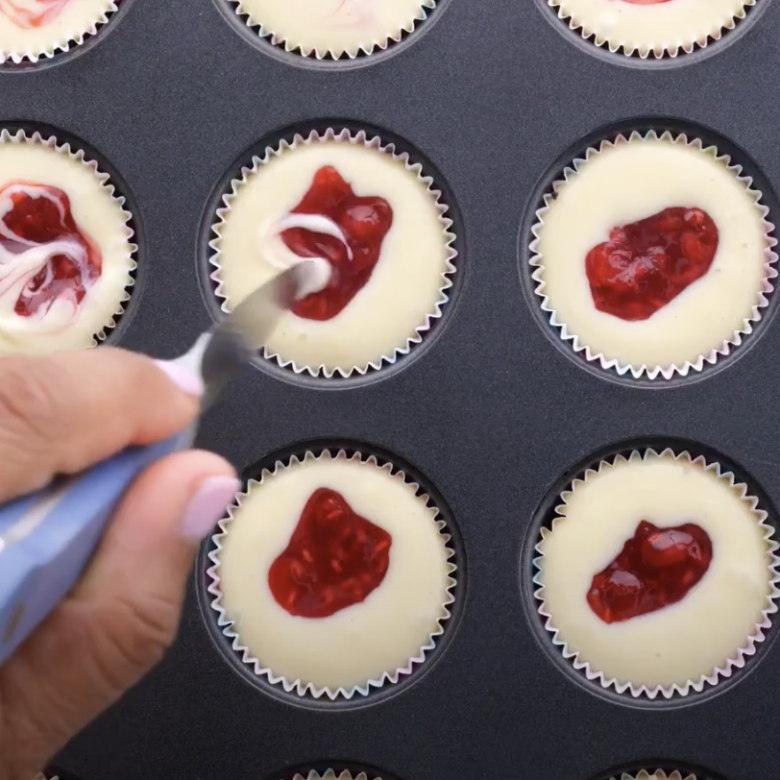 Raspberry sauce being swirled into cream cheese mixture in muffin pans.