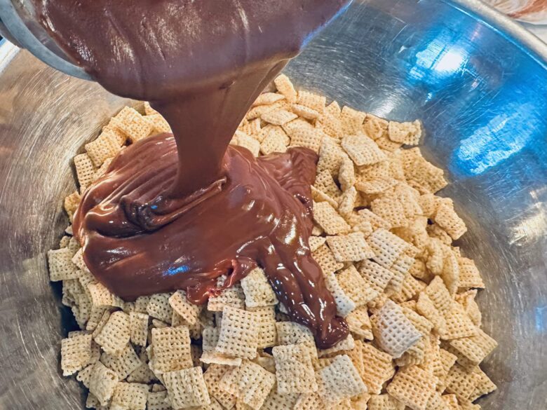 Melted chocolate mixture being poured onto Chex cereal in a large mixing bowl.