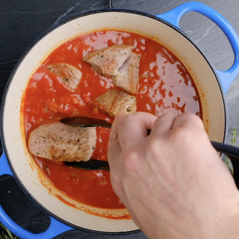 Seared pork being added to a Dutch oven with a tomato-based broth.