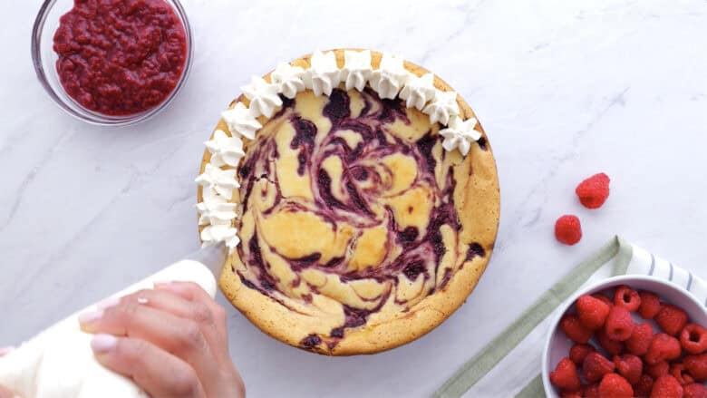 Whipped cream being piped on top of cooled raspberry cheesecake.