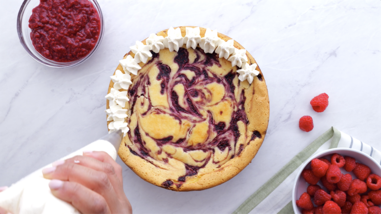 Whipped cream being piped on top of cooled raspberry cheesecake.