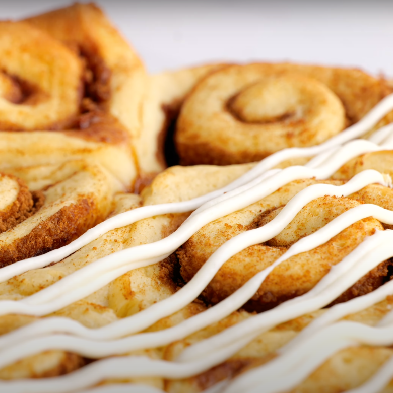 Freshly baked cinnamon rolls with cream cheese frosting being drizzled on top.